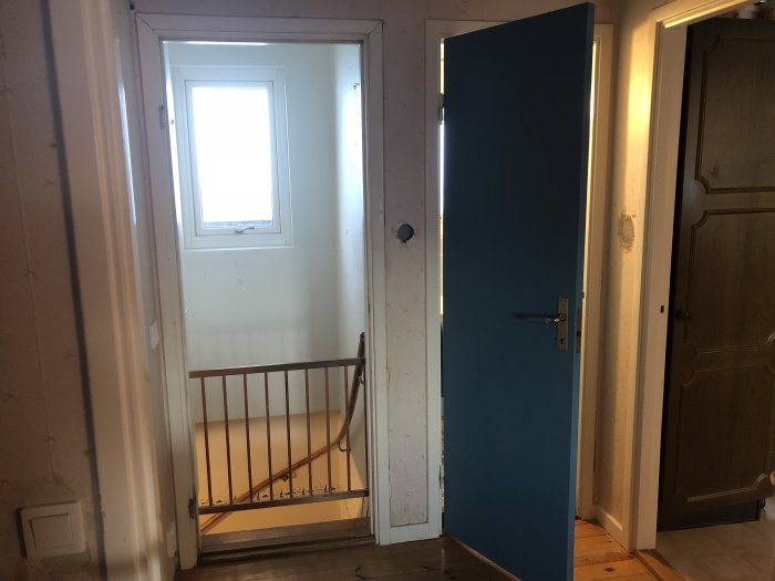 Vy från markplan över källartrappan med vit vägg och ett fönster, blå dörr öppen till vänster, och en stängd brun dörr.