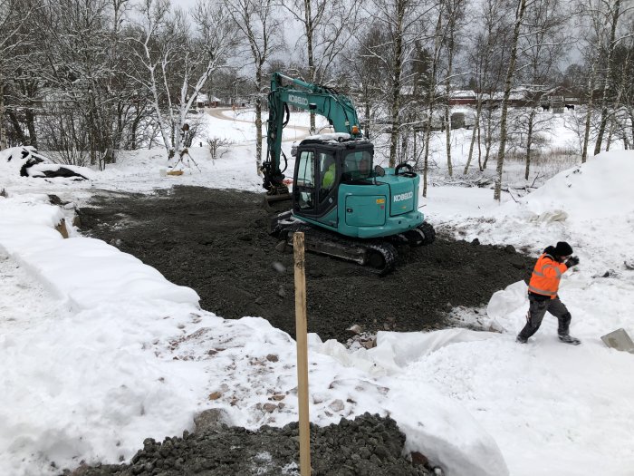 En person och en grävmaskin arbetar på en flisbädd vid ett vinterbygge.