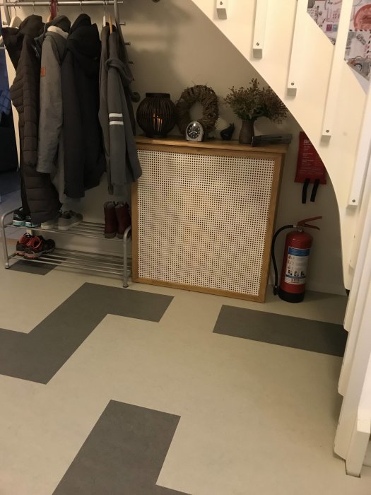 Hall med kalkvit vägg under trappa, jackor på klädhängare, skor och dekorativ radiator.