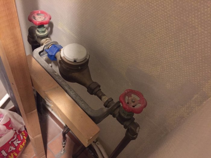 Vattenmätare stabiliserad med träregel mellan två röda ventiler mot en vägg.