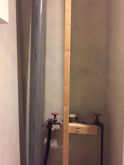 Vattenmätare stabiliserad med träregel mellan rör och vägg, inkluderar två rattar.