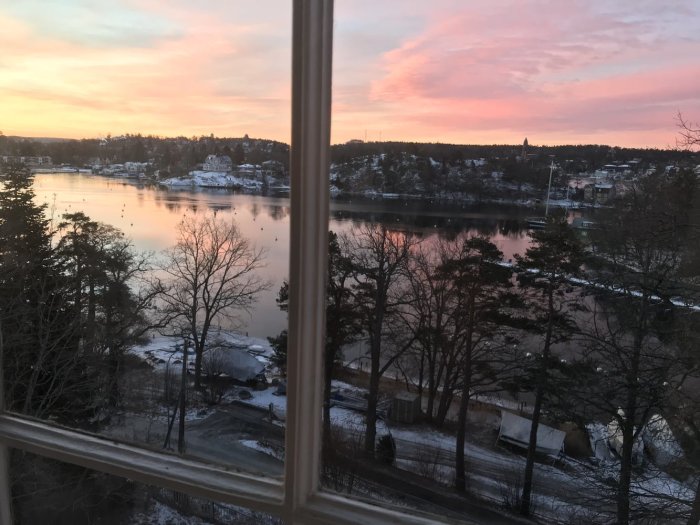 Utsikt från fönster i gammalt hus över vinterlandskap vid sjö i skymningen med rosa himmel.