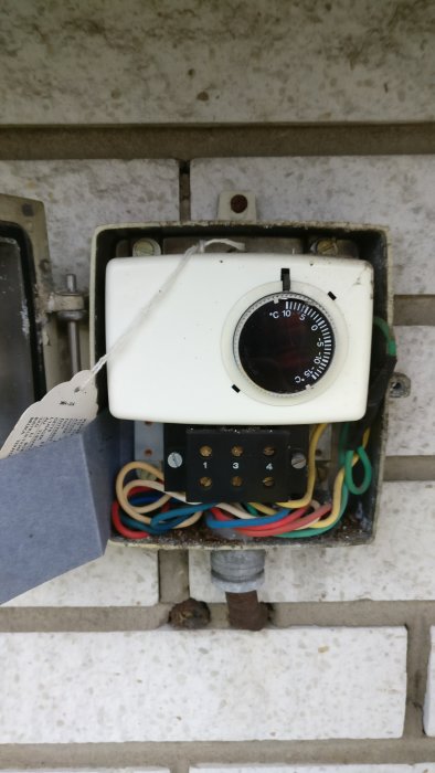 Gammal termostat med ratt och flera färgade kablar i ett öppnat elektriskt utrymme på en vägg.