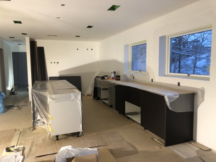 Köksrenovering pågår med omonterade svarta skåp och uppmätta väggar för bänkskivor, snöutsikt genom fönster.