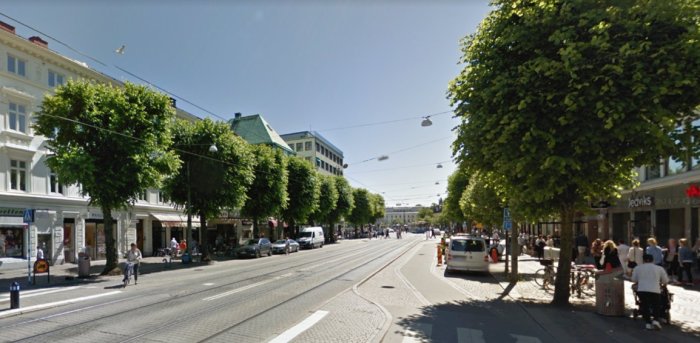 Stadsgata med spårvagnsspår, gående, cyklister och gröna träd på en solig dag.