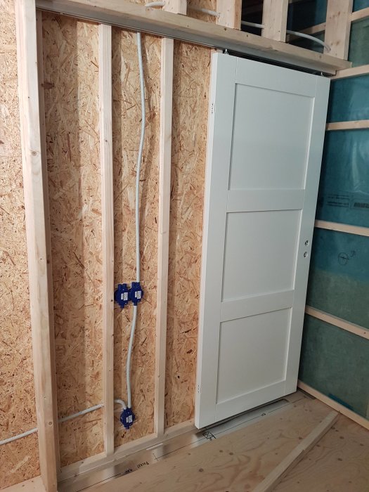 Installation av en vit skjutdörr in till en klädkammare med oskivade träväggar och elinstallationer synliga.