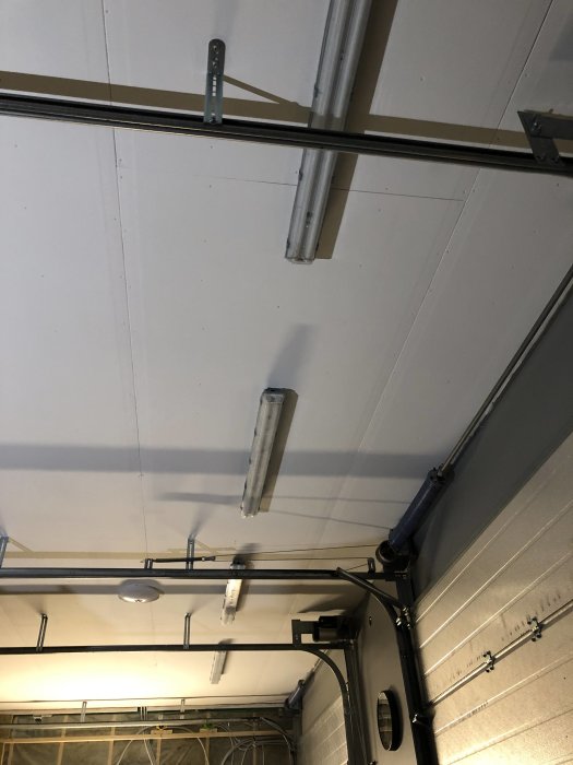 Lysrör och LED-armaturer monterade under ett garageportsspår nära taket, med synligt avstånd för att passa.