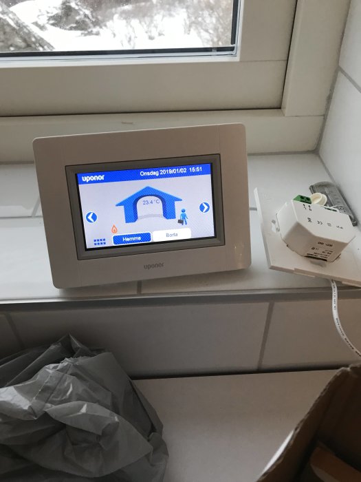 Uponor Smartix Wave manöverpanel för golvvärme med temperaturvisning 23.4 C och hem-borta läge mot ett fönster.
