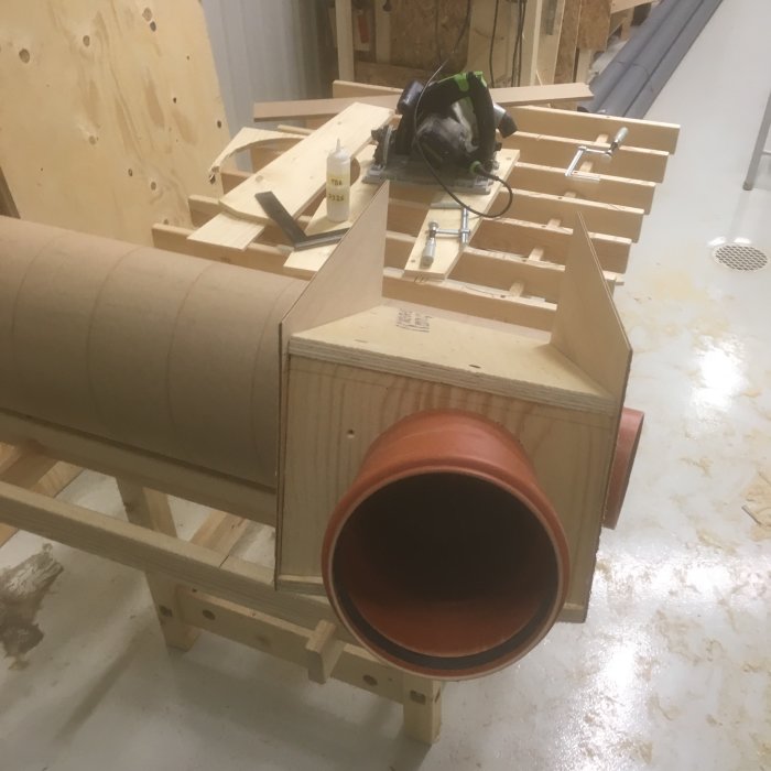 Delvis konstruerad träcyklon med en spåntratt av terrakottafärgade rör komponenter, omgiven av verktyg och träbitar på ett arbetsbord.