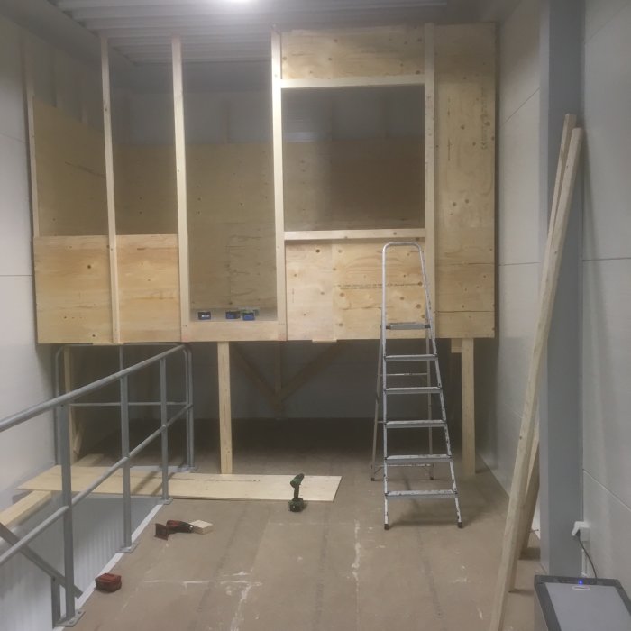 Påbörjad inredning av loft med trästommar och plywood, stege i förgrunden och verktyg på golvet.