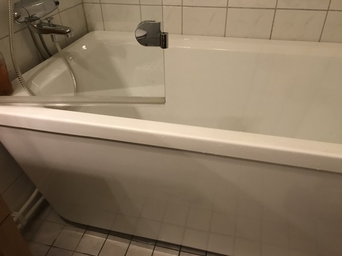 Vit hörnbadkar med obruten frontpanel i ett badrum med vita kakelväggar.