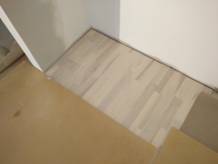Hörn av ett rum där ett nytt ljust trägolv möter äldre beige klinker.