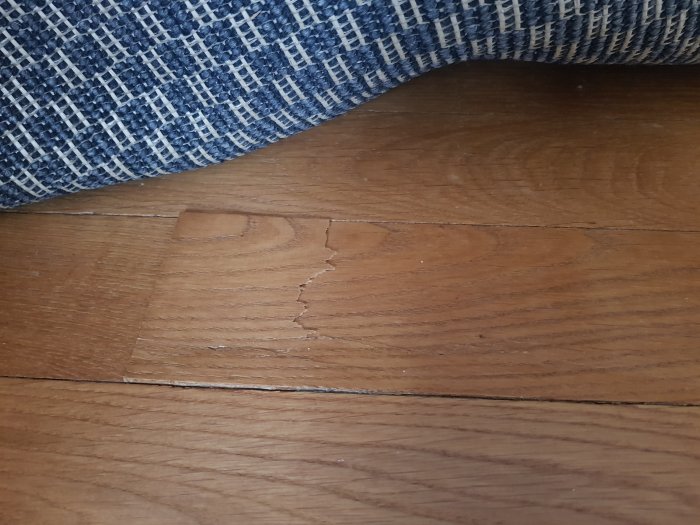 Trasigt parkettgolv med en knäckt och upphöjd stav nära en möbel med tygklädsel.