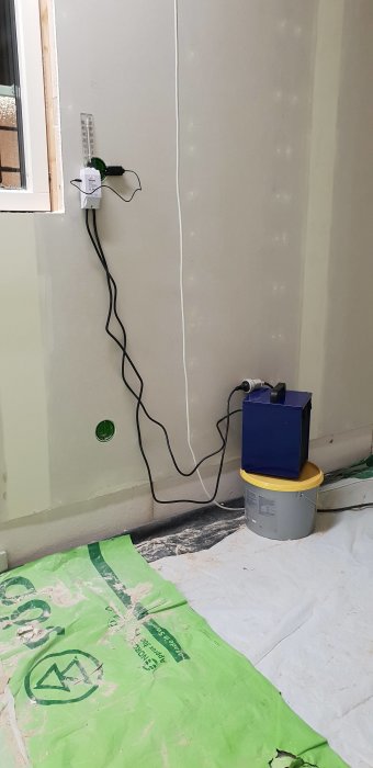 Eluttag med anslutna kablar och en blå värmefläkt på en gul hink i ett oisolerat garage.