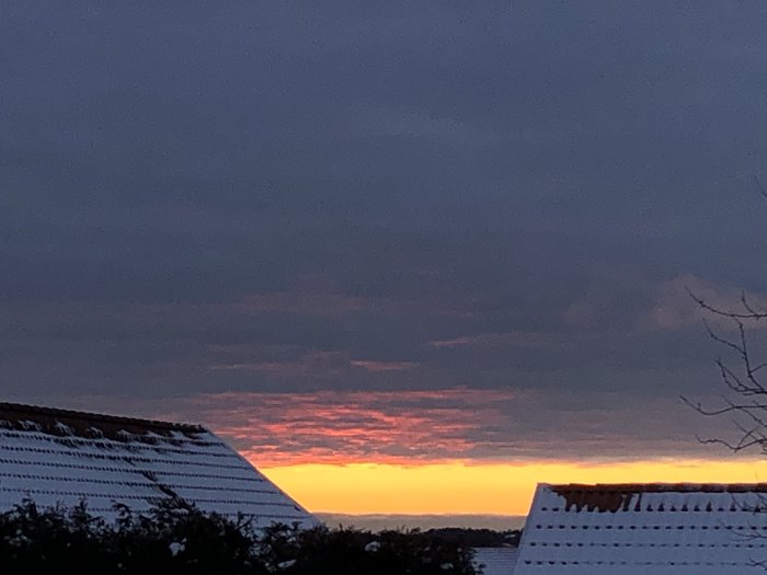 Tak med snö på grannens hus mot en dramatisk solnedgång med röda och gula nyanser.