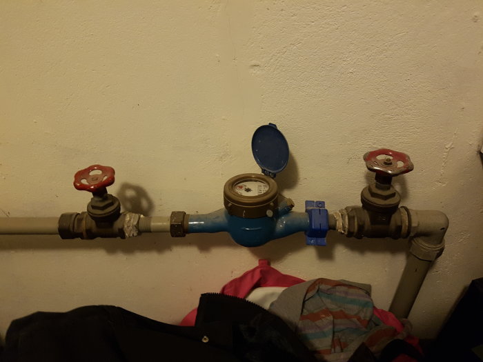 Vattenmätare med röda ventiler och blå fjärrmodul på vägg ovanför kläder.