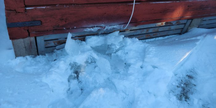Is och snö upphackad från marken mot en röd vägg, tecken på istillväxt vid ett hus.