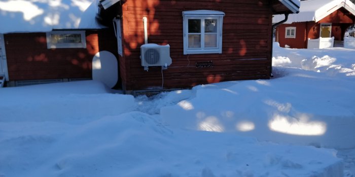 Luftvärmepump monterad på en snötäckt vägg av en röd stuga med isbildning nära marken.