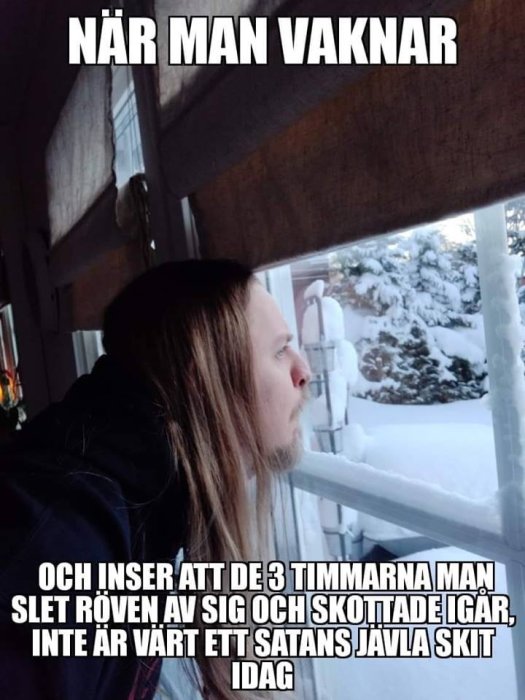 Person som tittar ut genom fönster på snöig trädgård, synbart besviken eller frustrerad.