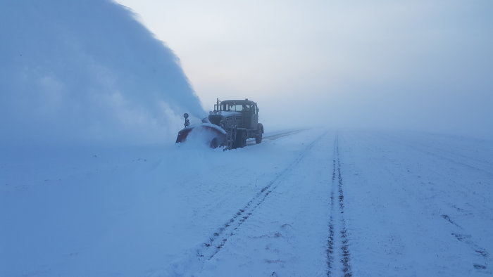 Traktor med snöslunga i arbete på en snötäckt väg i disigt vinterlandskap.