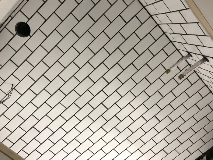 Vit kaklad vägg med mörk fog som visar framträdande sprickor i vissa kakelplattor och synliga rörgenomföringar.