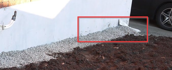 Grundmur med grus och markisolering vid husvägg, synlig del av stuprör i bakgrunden.