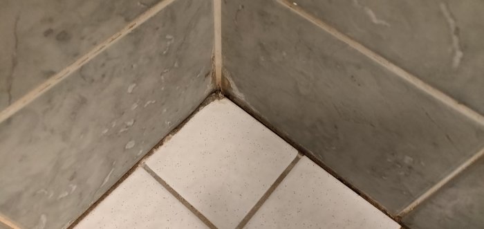 Hörnet av en dusch med grå väggkakel och vit golvkakel där fogmassan är skadad och saknas i skarven.
