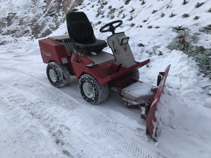 Röd hydraulisk driv- och lyftutrustad plogmaskin på ett snöigt underlag med ett snötäckt landskap i bakgrunden.