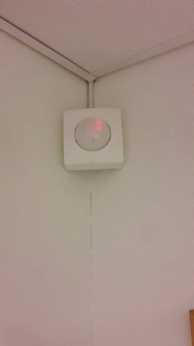 Vit rörelsevakt monterad i takhörn med rött ljus som indikerar aktivering eller fel.