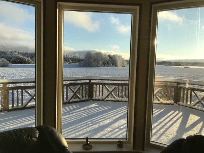 Utsikt från ett fönster över snötäckt landskap och en trädäckad balkong i solljus.