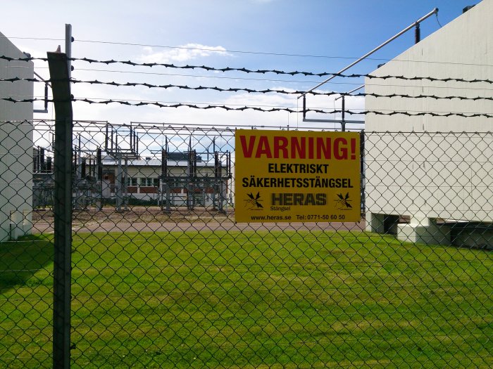 Stängsel med varningsskylt för elektriskt säkerhetsstängsel framför industribyggnad.