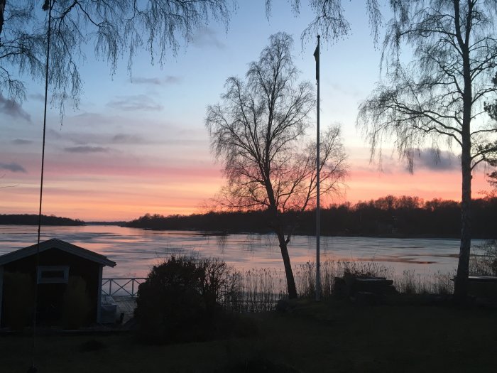 Solnedgång med färggrann himmel över en isbelagd sjö med silhuetter av träd och en liten stuga.