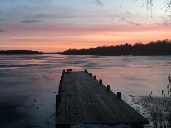 En träbrygga som leder ut till en frusen sjö vid solnedgång med rosa och blå himmel.