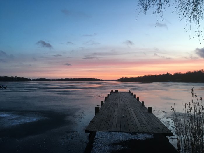 Brygga som leder ut till en frusen sjö vid solnedgång med rosa och blå himmel.