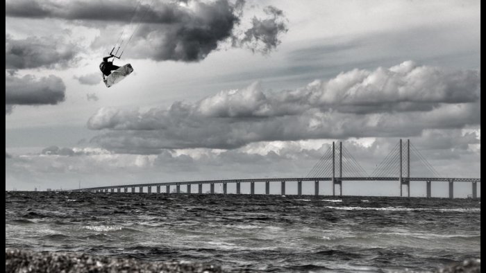 Kitesurfare i luften över havet med en lång bro i bakgrunden under en molnig himmel.