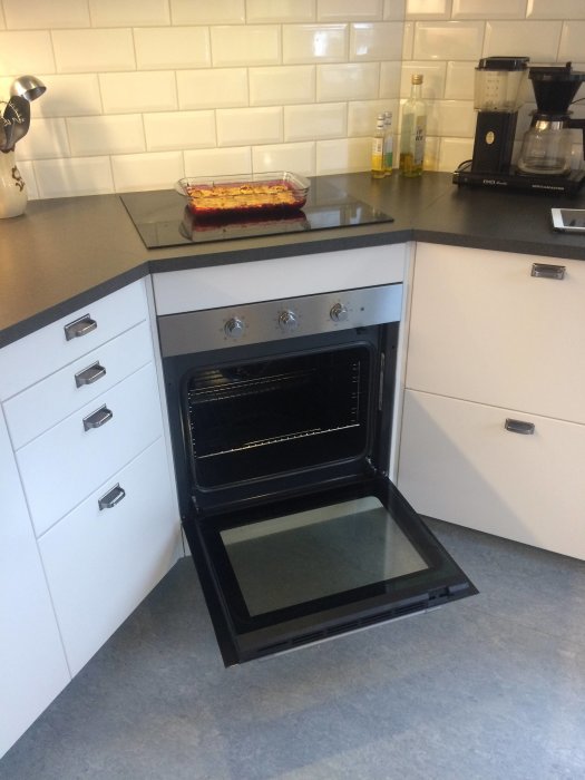 Öppen ugnslucka i kök med lasagneform på bänkskivan och köksutrustning i bakgrunden.