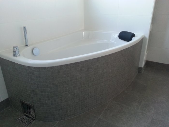 Hörnbadkar klätt med grå mosaik och fastlimmade lux-elementskivor i badrum.