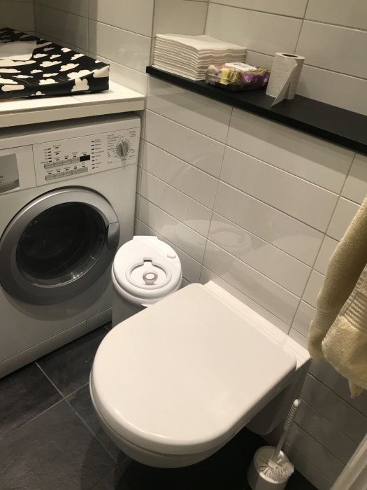 Badrum med vägghängd WC intill tvättmaskin, vita kaklade väggar och gråa klinkers på golvet.