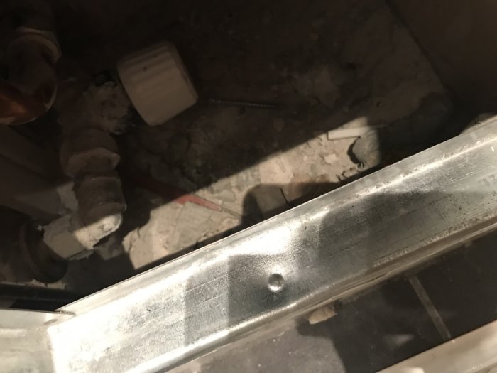 Oskyddad röranslutning och betonggolv synligt bakom en vägghängd toaletts öppna installationsyta.