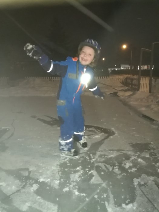 Ett leende barn i vinterkläder åker skridskor på en hemmagjord isbana i trädgården på kvällen.