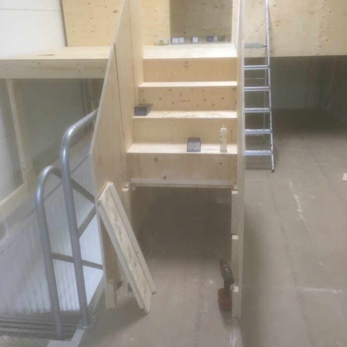 Ofärdig trätrappa under konstruktion med oskruvade steg och täckta sidor, verktyg och byggmaterial synligt.