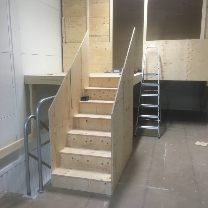 Halvfärdig trappa med synliga skruvar och täckta räcken av plywood i ett pågående byggprojekt.