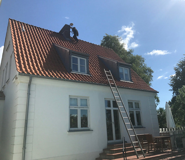 Sotare i arbete på ett tegeltak på en vit tvåvåningshus med en stege som leder upp till skorstenen.