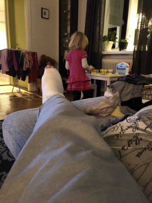 Person med gipsad fot framför en liten flicka som leker i ett vardagsrum.