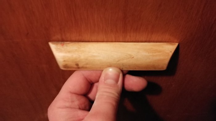 Nött trähandtag från ett 50-tals kök, hållet av en hand mot brun skåpsdörr, möjlig kantlist synlig.