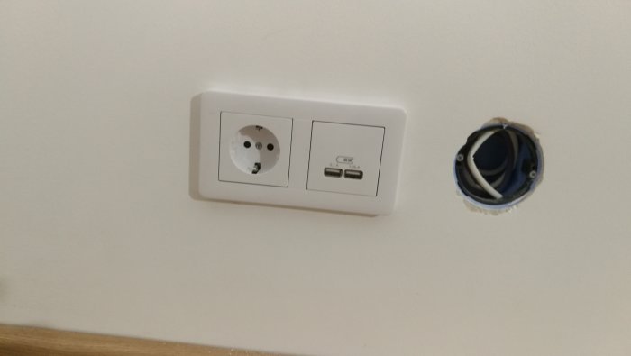 Vägguttag med en vanlig kontakt och dubbla USB-laddningsportar intill ett omonterat elutrymme.