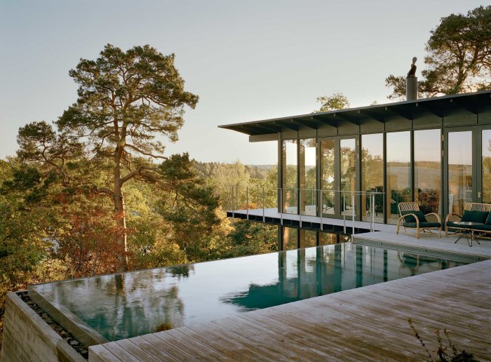 Modern hus med infinitypool och en altan av trä, omgiven av natur och ett ljuskärt interiört utrymme synligt genom stora fönster.