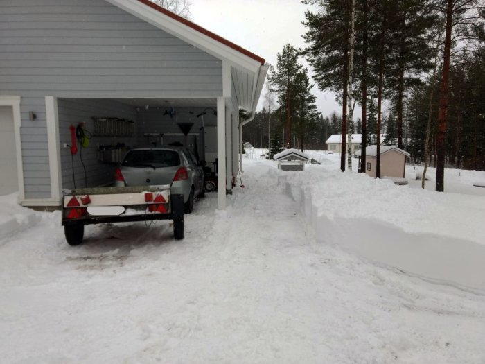 Snöig uppfart med upplogad snövägg och släpvagn framför garage med bil inuti.