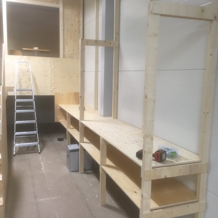 Nybyggd lagerhylla i trä med övre och nedre plan och en stege, i ett ljust rum under konstruktion.