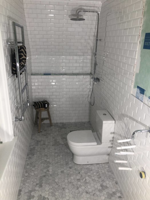 Renoverat badrum med vita subway-kakelväggar, stenmosaikgolv, monterad toalett och handdukstork.